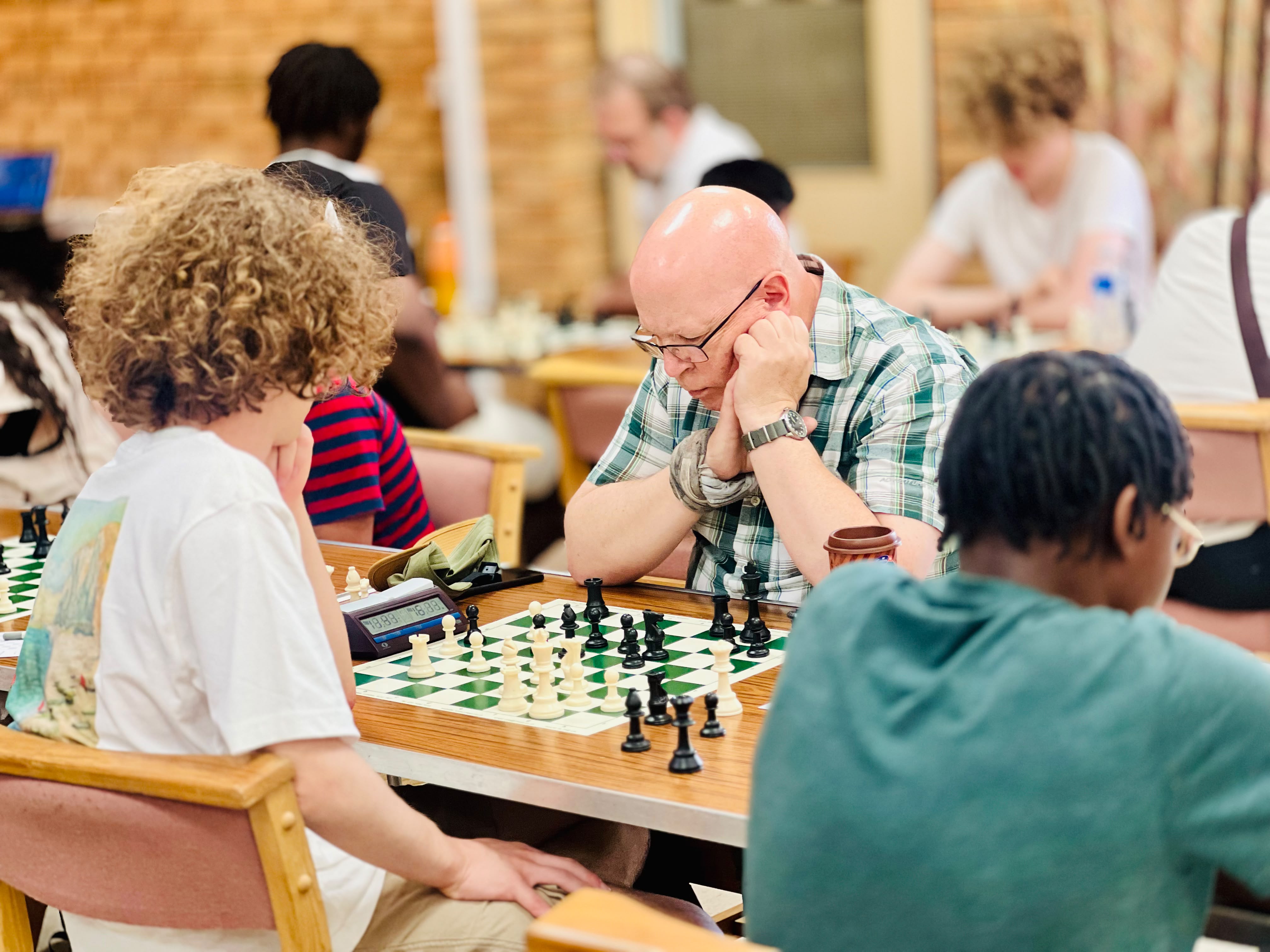 Adult player deep in concentration facing junior players across multiple chess boards in a busy tournament hall