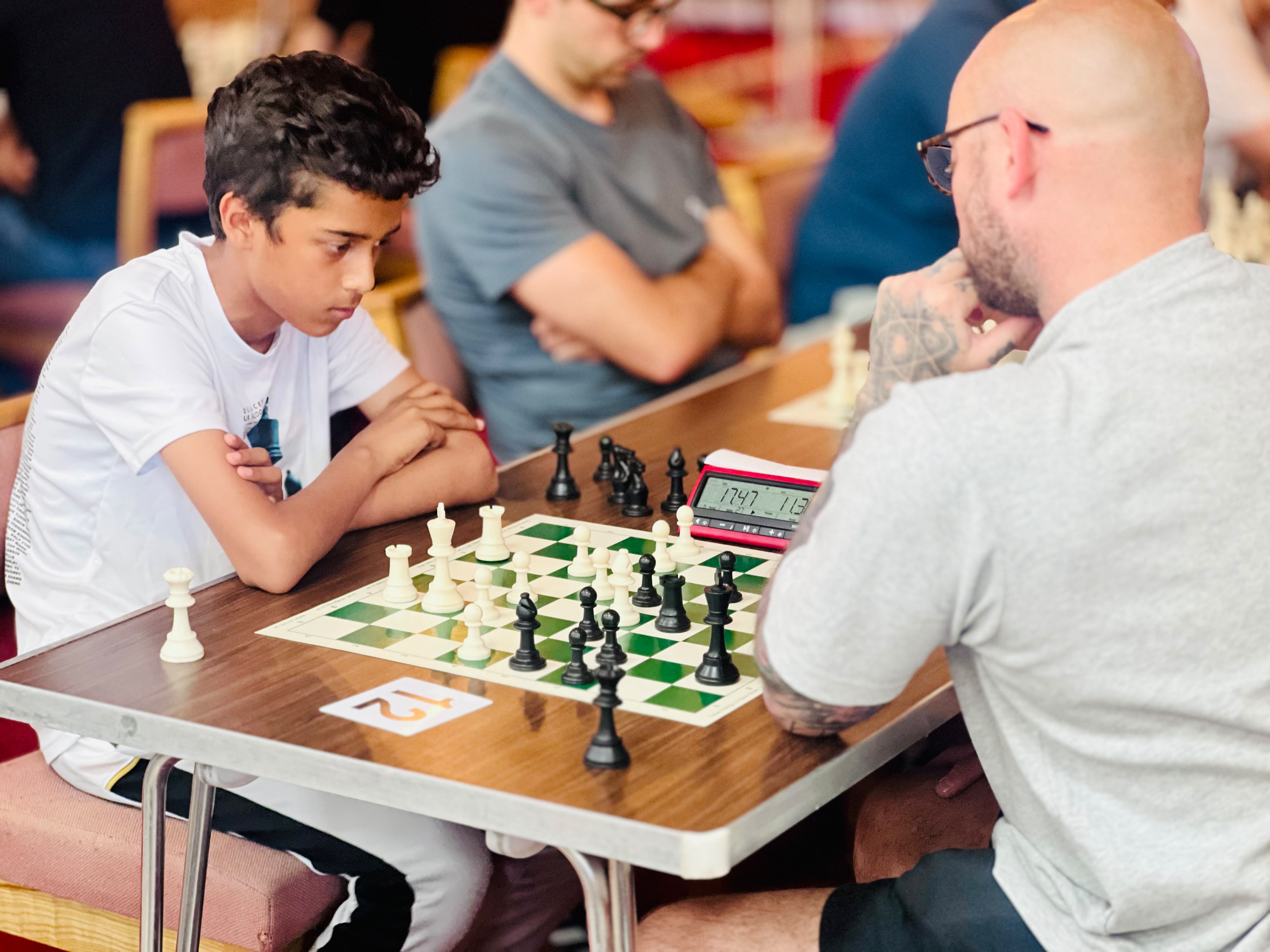 Teenage player with arms crossed studying the board intently against an adult opponent, chess clock visible